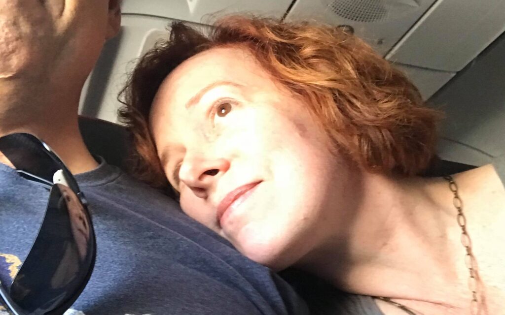 Edie White rests her right cheek against Jeremy White's chest as she peers out of an unseen airplane window.