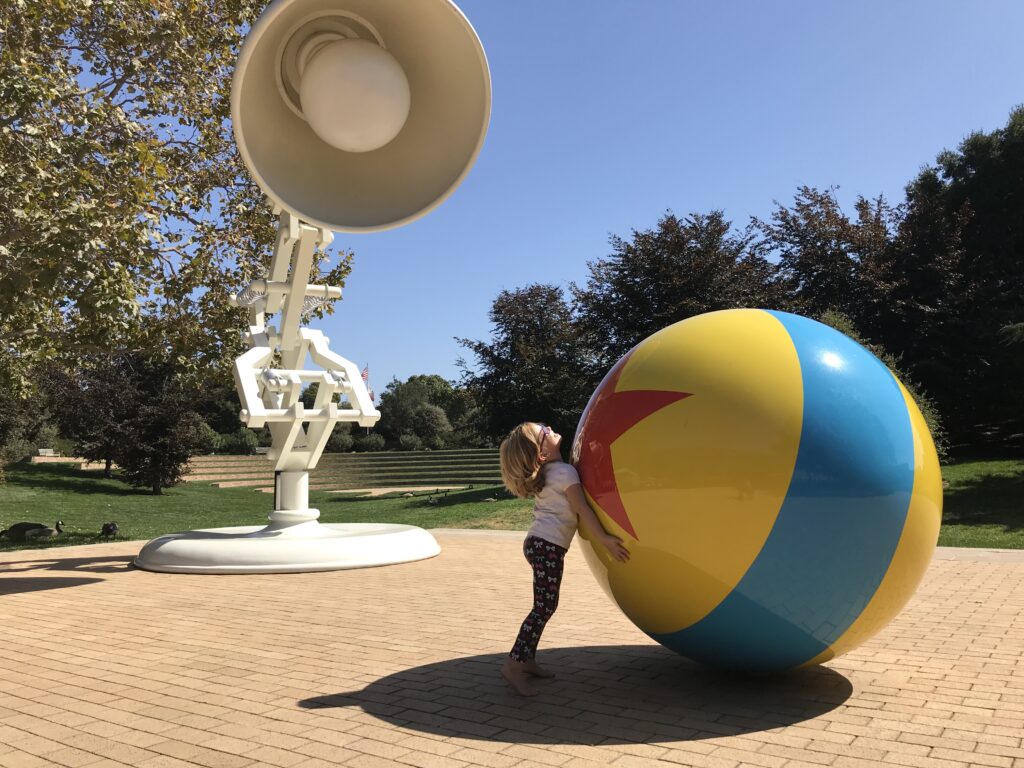 A young girl attempts to move a large model of the Pixar ball in a bricked courtyard overseen by it's companion Luxo.